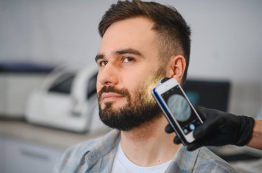 Özel bir el bilgisayarı kullanarak kafa derisi, saç ve sakal durumu değerlendirmesi için trikoloğa giden hasta.