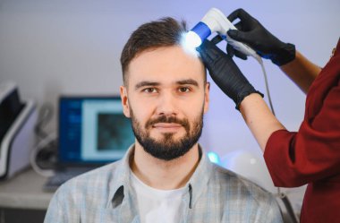 Genç adam saç dökülmesi sorunlarıyla karşı karşıya. Modern bir klinikte kafa derisi muayenesi yaptırıyor, profesyonel bakım görüyor.