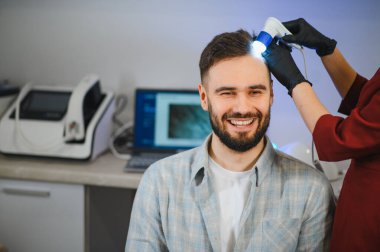 Mutlu bir adam, saç dökülmesi ve dermatoloji için özel bir cihaz kullanarak trikoloğun birinden profesyonel kafa derisi muayenesi alıyor.