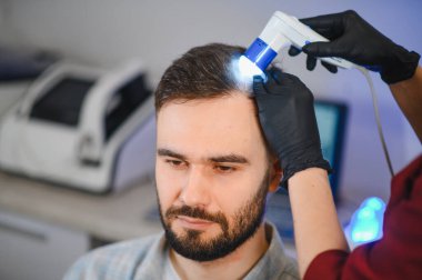 Kafa derisi ve saç folikülleri muayenesinden geçen bir erkek hasta. Klinikteki bir trikoloğun profesyonel dermatoskopuyla incelemesi.