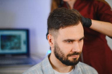 Genç bir adam, modern bir klinikte kafa derisini ve saçını inceleyen profesyonel bir trikoloğa sahip.