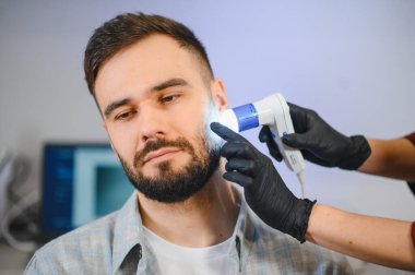 Uzman trikoloğum hastanın sakalını ve cildini cilt bakımı ve cilt bakımı için özel ekipmanla inceliyor.