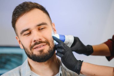 Genç adam, modern bir klinikteki bir trikoloğun özel bir cihazıyla detaylı bir sakal ve deri analizi yaptırıyor.
