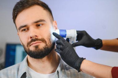 Genç bir adam, bir uzman tarafından modern bir dermatoskop cihazı kullanılarak sakalına detaylı bir deri ve saç folikül kontrolü yaptırıyor.