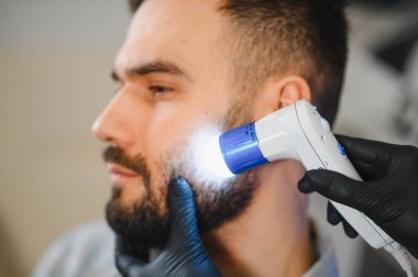 Dermatolog, cilt bakımı sırasında erkek bir hastanın deri ve sakalını incelemek için dermatoskop kullanıyor.