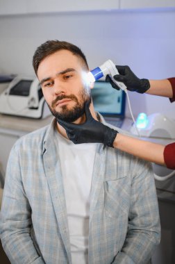 Trikoloğum, bir klinikte saç kaybı ve deri sağlığı için dermatoskopla erkek hastanın kafa ve sakalını inceliyor.