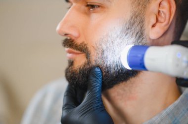 Bir tricholog tarafından sakal ve yüz bakımı yapılan erkek hasta özel bir el bilgisayarı kullanıyor.