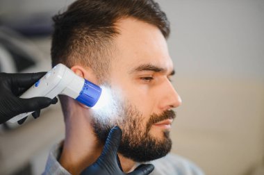 Dermatolog hastanın sakalını ve cildini dermatoskopla inceliyor. Profesyonel sağlık hizmeti sağlayıcısı cilt analizi yapıyor