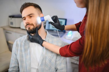 Trikoloğum, bir adamın sakalını ve yüzünü klinik ortamında özel bir aletle inceliyor. Deri ve saç hastalıklarını teşhis ediyor.