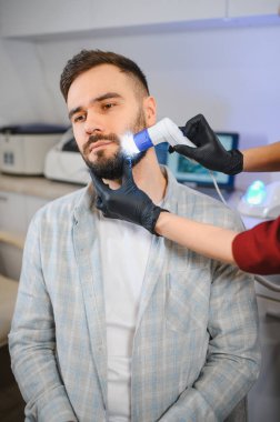 Genç bir adam, özel bir el bilgisayarı kullanarak sakalını ve derisini muayene etmek için bir dermatoloğu ziyaret ediyor.