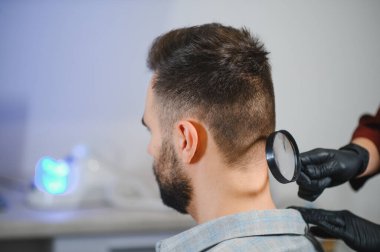 Siyah eldivenli doktor bir adamın boynunu büyüteçle inceliyor, benleri ve deri bozukluklarını kontrol ediyor.