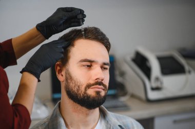 Sakallı bir adam saç yenileme prosedüründen geçiyor. Eldivenli bir doktor tarafından kafa derisine serum uygulanıyor.