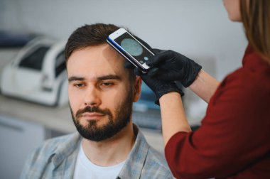 Trikoloğum, saç kaybı, kellik teşhisi ve tedavi için telefon bağlantılı bir dermatoskop kullanarak erkek hastanın kafa derisini inceliyor.