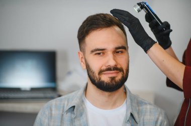 Trikoloğum, saç kaybı teşhisi ve tedavi planlaması için dermatoskop kullanan bir erkek hastanın kafa derisini inceliyor.