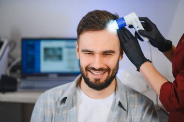 Trikoloğa giderken gülümseyen genç bir adam saç dökülmesi ve bakım için dermatoskopla kafa derisini incelettiriyor.