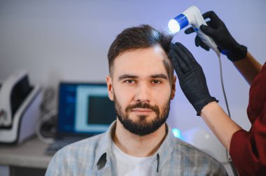 Genç bir adam kafa derisi muayenesi için bir trikoloğu ziyaret ediyor, saç problemleri için teşhis ve tedavi görüyor.