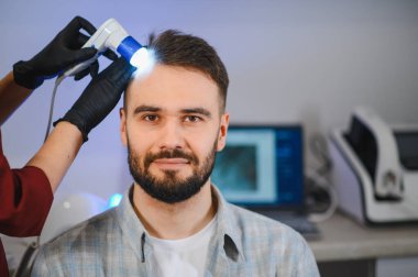 Trikoloji muayenesinden geçen bir adam, saç sağlığı için kafa derisini modern bir tıbbi aletle kontrol eden bir uzman.