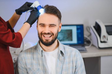 Profesyonel bir trikoskopi sırasında gülümseyen genç adam, klinikte saç sağlığı ve kafa derisi durumunu kontrol ediyor.