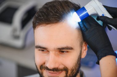 Siyah eldivenli trikoloğum gülümseyen bir adamın kafa derisini modern bir dermatoskopla inceliyor, saç sağlığını kontrol ediyor.