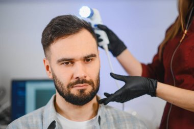 Trikoloğun teki klinikte özel bir aletle kafa derisini ve saçını incelerken erkek hasta öylece oturuyor.