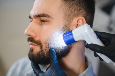 Trikoloğum bir adamın sakalını ve altında yatan deriyi muayene ederken özel ışıklandırılmış tıbbi bir cihaz kullanıyor.