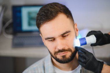 Tıbbi klinikte trikoloğum saç dökülmesi teşhisi için dermatoskopla erkek hastanın kafa derisini ve saç foliküllerini inceliyor.