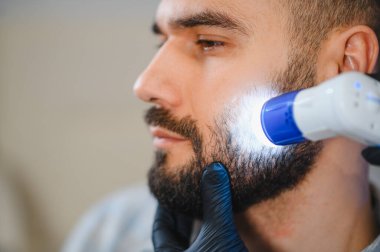 Erkek hasta, dermatoloji kliniğinde sakalının ve cildinin trikoskopi muayenesinden geçiyor.