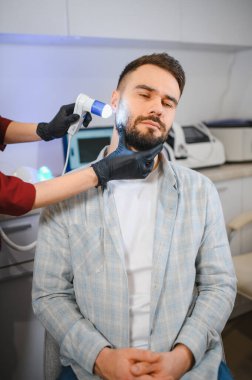 Erkek hasta trikoloji kliniğinde uzman biri tarafından dermatoskopla sakal ve cilt muayenesi yaptırıyor.