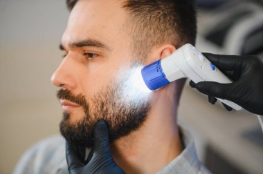 Trikoloğum bir adamın yüz kıllarını ve cilt hastalığını dermatoskopla kontrol ediyor.