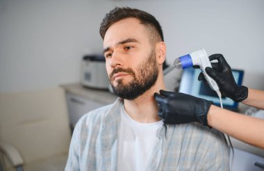 Genç bir erkek hasta trikoloji kliniğinde özel bir tıbbi aletle kafa derisi ve saç analizi yaptırıyor.