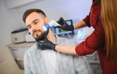 Trikoloğum, klinikte özel bir dermatoskop kullanarak erkek bir hastanın sakalı ve yüz derisi üzerinde teşhis incelemesi yapıyor.