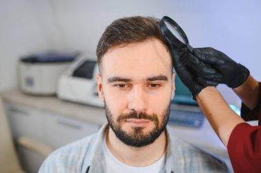 Siyah eldivenli trikoloğum saç kaybını tespit etmek için bir büyüteç kullanarak genç bir hastanın kafa derisini inceliyor.