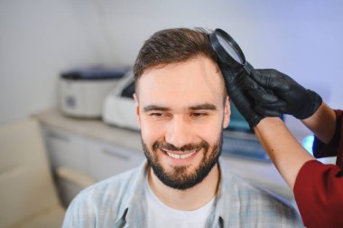 Bir trikoloğu ziyaret eden gülümseyen adam, saç ve kafa derisini klinikte büyüteç kullanarak incelettiriyor.