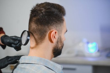 Doktor dermatoloji kanseri taraması sırasında bir büyüteç kullanarak bir adamın derisinde leke ve anormallikleri kontrol ediyor.