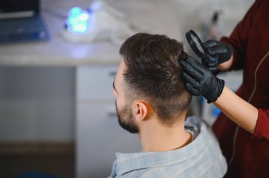 Erkek hastanın kafa derisini ve saçını trikoloji ya da saç kaybı muayenesi için bir klinikte büyüteçle inceleyen eldiven takan bir dermatolog.