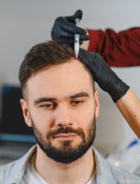 Genç adam trikoloğundan büyüme faktörü enjeksiyonu alıyor, saç dökülmesini ele alıyor ve bir klinik ortamında kafa derisi sağlığını destekliyor.