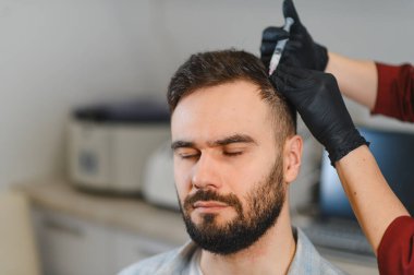 Trikoloğun biri saç dökülmesini tedavi etmek için kafa derisine mezoterapi yaparken hasta gözleri kapalı oturuyor.