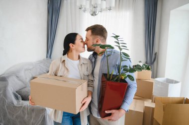 Mutlu genç çift öpüşüyor, taşınma kutuları ve bitkileri tutuyor, yeni daireye taşınmayı kutluyorlar.