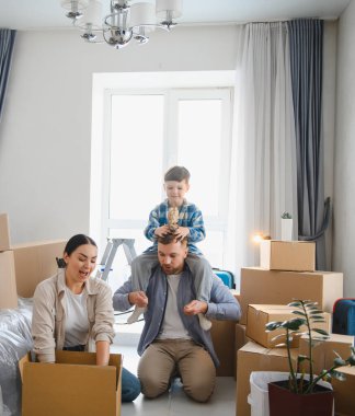 Mutlu bir aile yeni bir eve taşınıyor, karton kutuları açıyor ve babasının omuzlarında bir çocuk var.