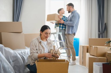 Mutlu bir aile yeni bir eve taşınıyor. Anne, karton kutularla çevrili eşyaları yerleştirirken baba ve oğul bir kutuyu dikkatlice tutuyor.