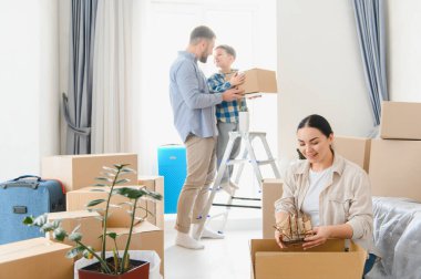 Mutlu bir aile yeni bir eve taşınıyor, karton kutuları açıyor, ev eşyaları ve kişisel eşyalarla birlikte.