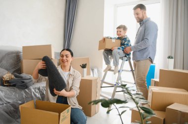 Genç aile yeni evine taşınmayı kutluyor, kutuları açıyor ve gülümseyen ve parlak iç mekanlarda takım çalışması yapan eşyalar düzenliyor.