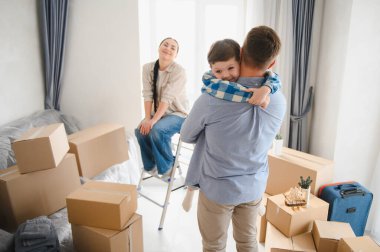 Aile yeni eve taşınmamızı kutluyor. Ebeveynler ve çocuklar karton kutularla sarılıyorlar. Mutlu hissediyorlar.