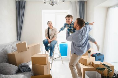 Mutlu bir aile yeni bir eve taşınıyor, baba oğluyla oynuyor, anne merdivende oturuyor karton kutuların arasında gülümsüyor.