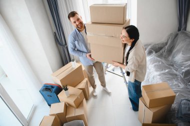 Karton kutuları taşıyan genç ve gülümseyen bir çift paketlenmiş eşyalarla çevrili yeni evlerine taşınıyorlar.