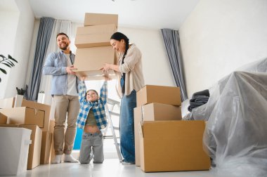 Genç bir aile taşınma kutularını parlak bir oturma odasına yerleştiriyor. Ebeveynler ve küçük çocuklar yeni bir başlangıç ve takım çalışması sırasında gülümsüyor.