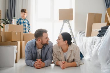 Genç bir çift ve çocuk yeni evlerinin zemininde dinleniyor, kutuları taşıyarak, taşınmayı kutluyorlar.