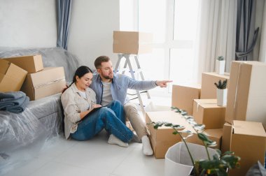 Karton kutuların arasında oturmuş, yeni dairelerinin tasarım fikirlerini tartışan mutlu çift.