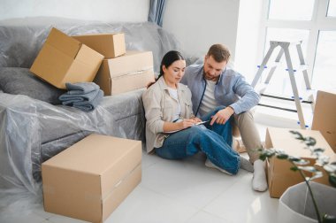 Bir çift yerde oturuyor, taşınma ve paketleme için kontrol listesini inceliyor, taşınan kutular ve örtülü mobilyalar ile çevrili.