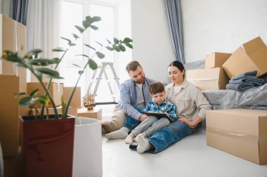 Çocuklu bir aile yeni evin zemininde oturuyor, taşındıktan sonra karton kutuların arasında tabletle rahatlıyor.